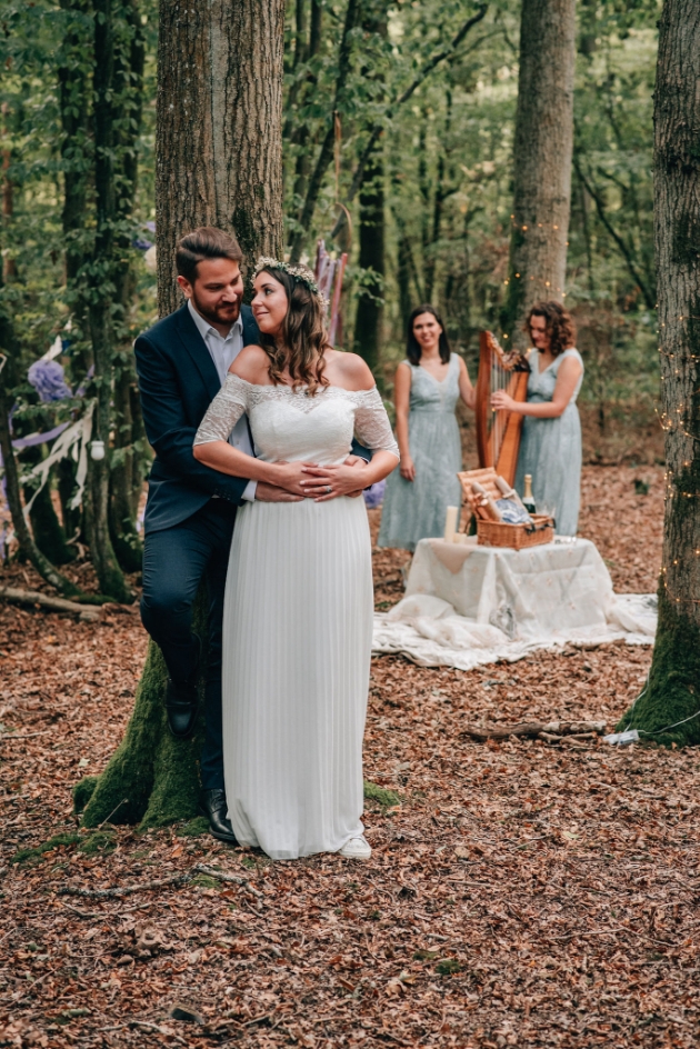 Bride and groom leaning against a tree in the woods. 2 of harps can be seen in the background