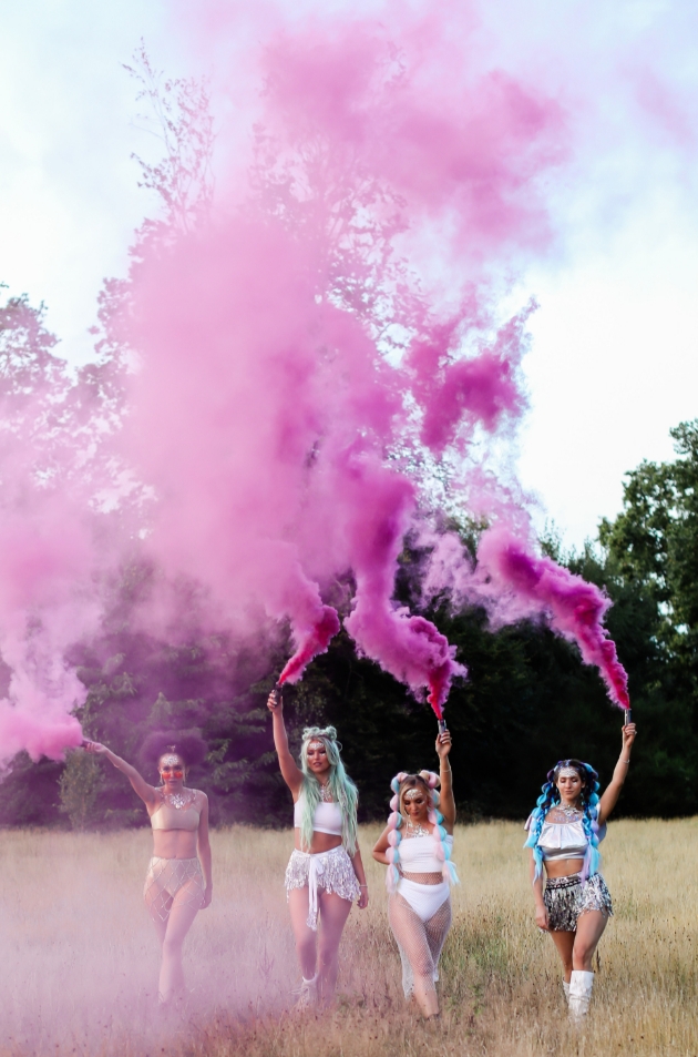 four women dressed in festival outfits walking in field with pink smoke bomb