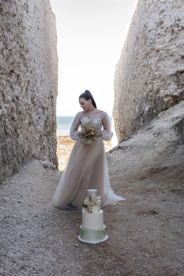 Model on a beach in Kent wearing boho dress holding bouquet standing behind a cake