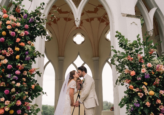 wedding couple kissing under wedding arch in garden covered in flowers