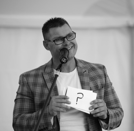 matty in a checked suit holding magic cards speaking in a mic