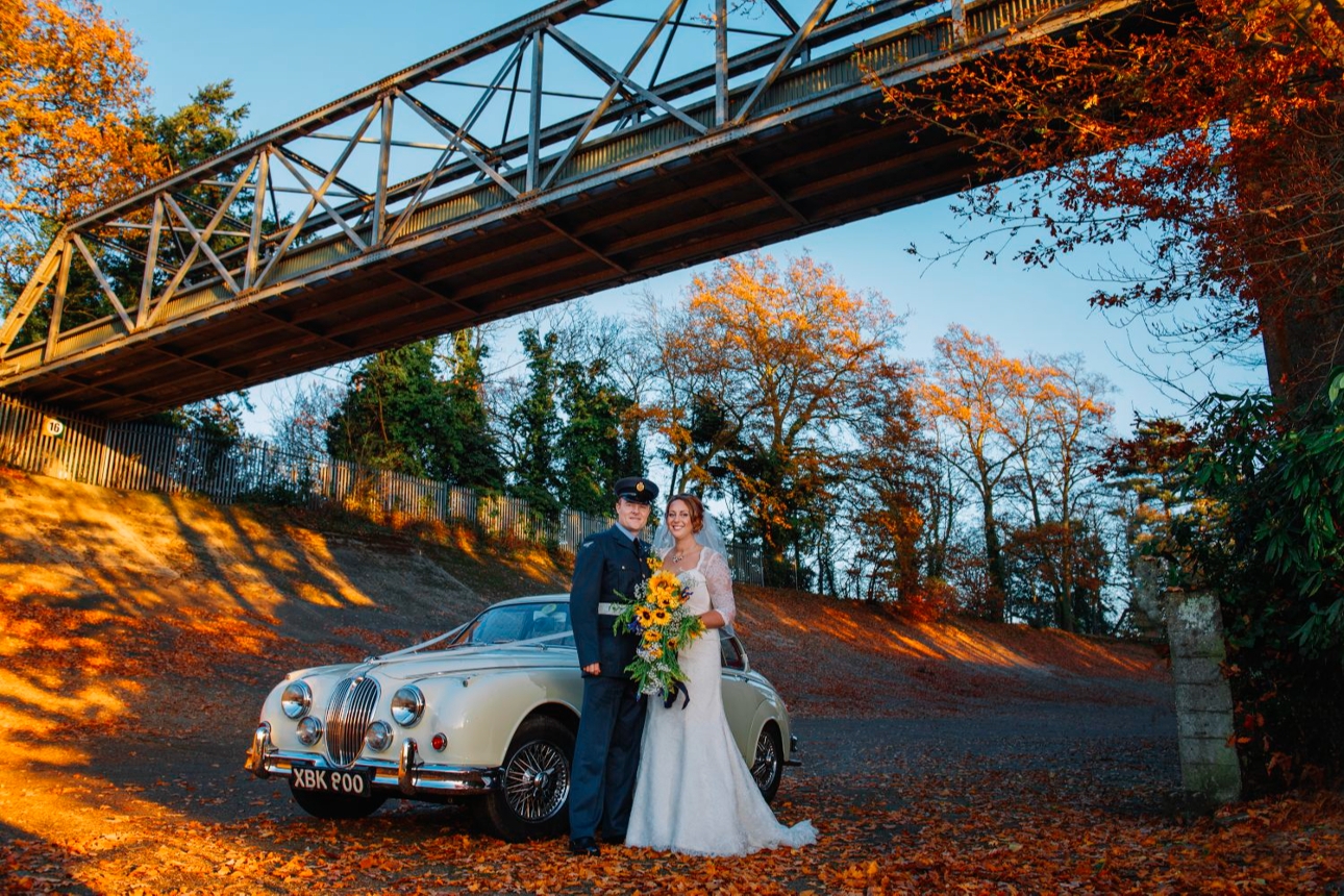 bride and groom standing in front of wedding car on autumnal day 