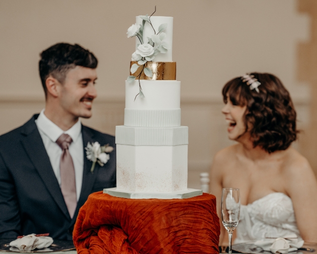 bride and groom smile at each other in background with wedding cake in foreground