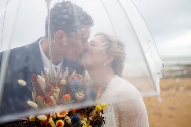 bride and groom kissing beneath an umbrella in the rain