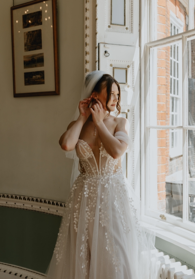 bride stood in front of a big window putting an earring in