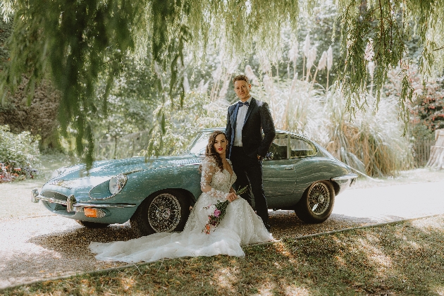 A bride and groom standing in front of a green car below a tree
